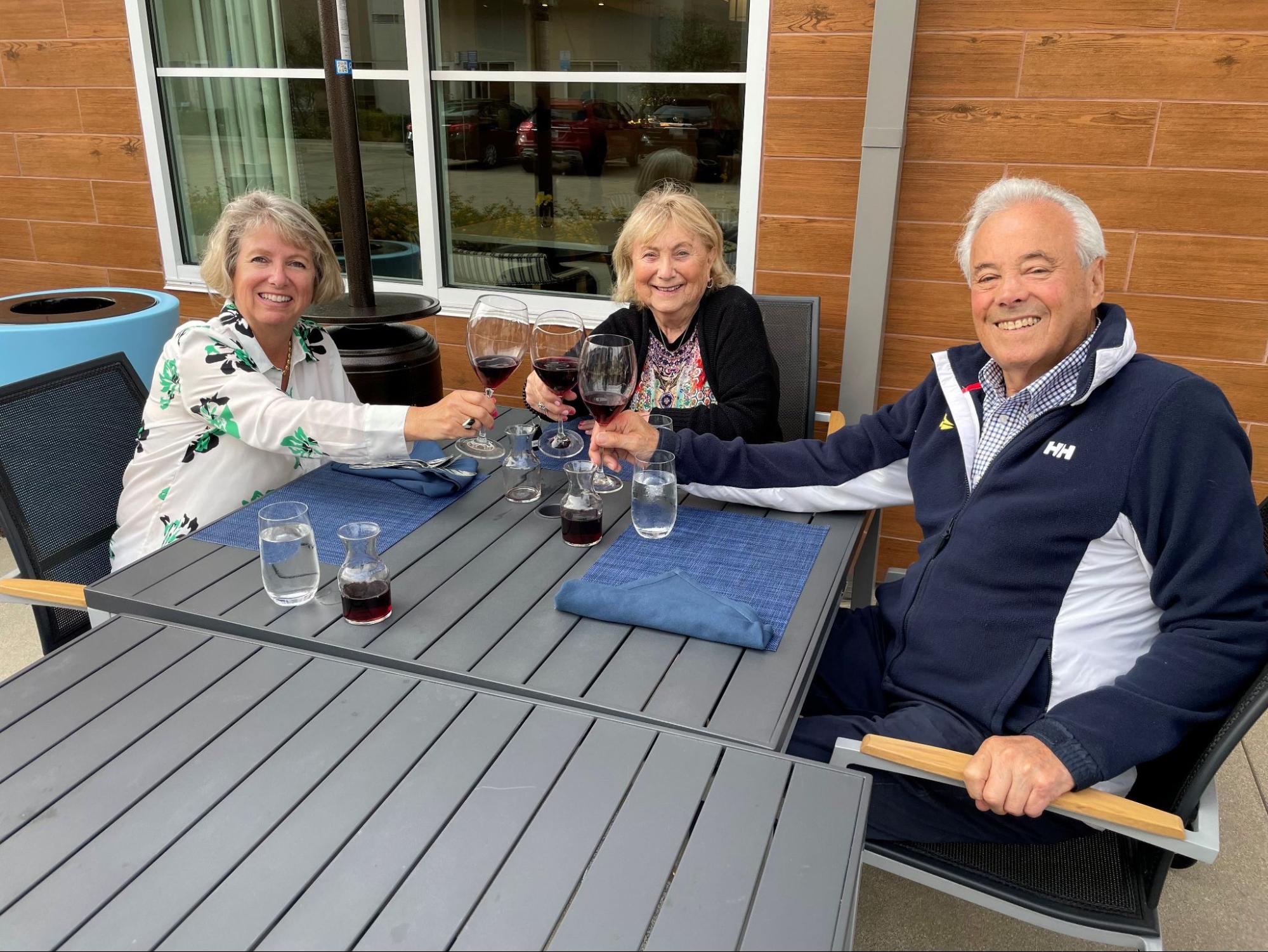 Three seniors sitting at a table with glasses of red wine, smiling for a photo.