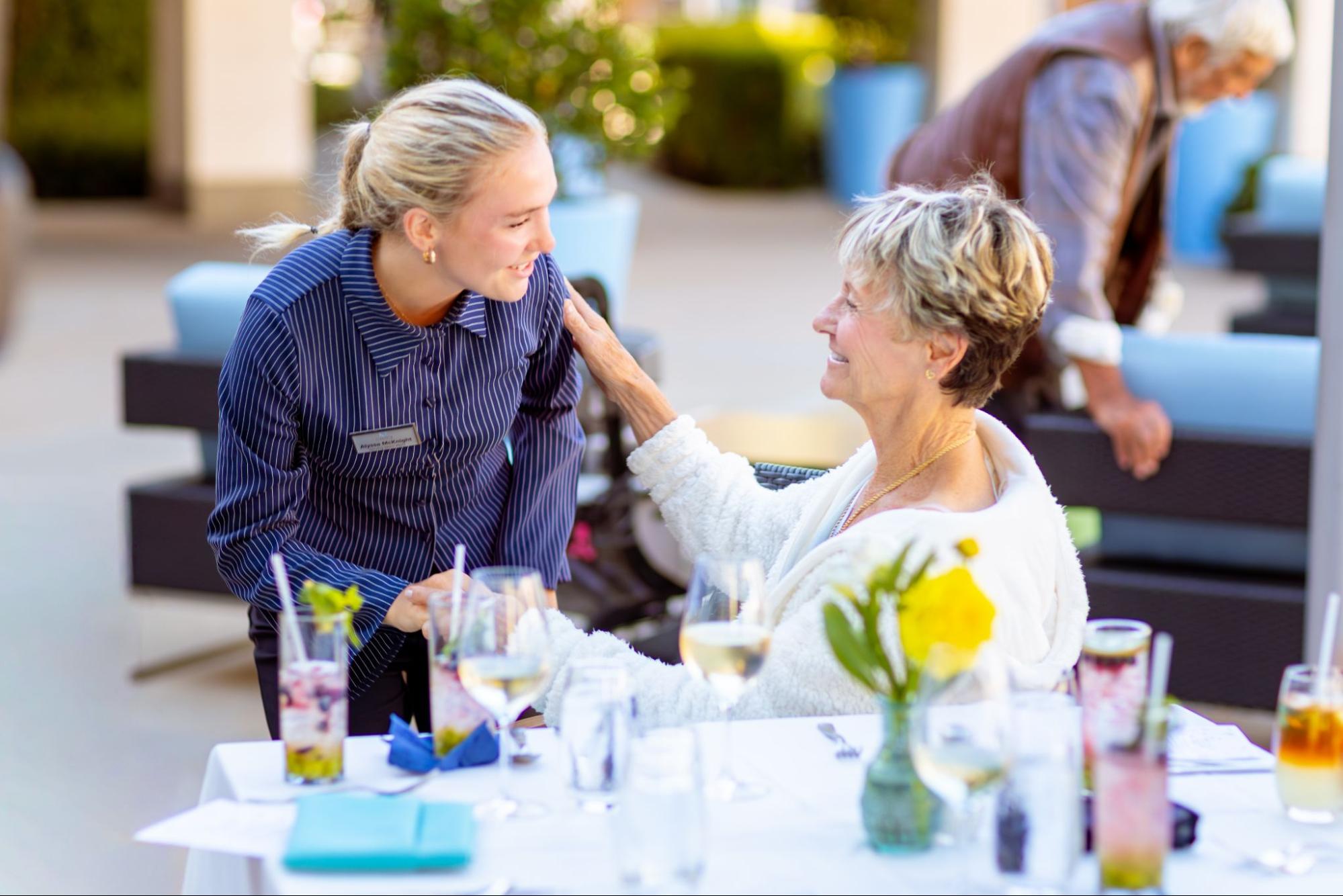 A young woman leaning down, smiling at an elderly woman sitting at an outdoor dining table.