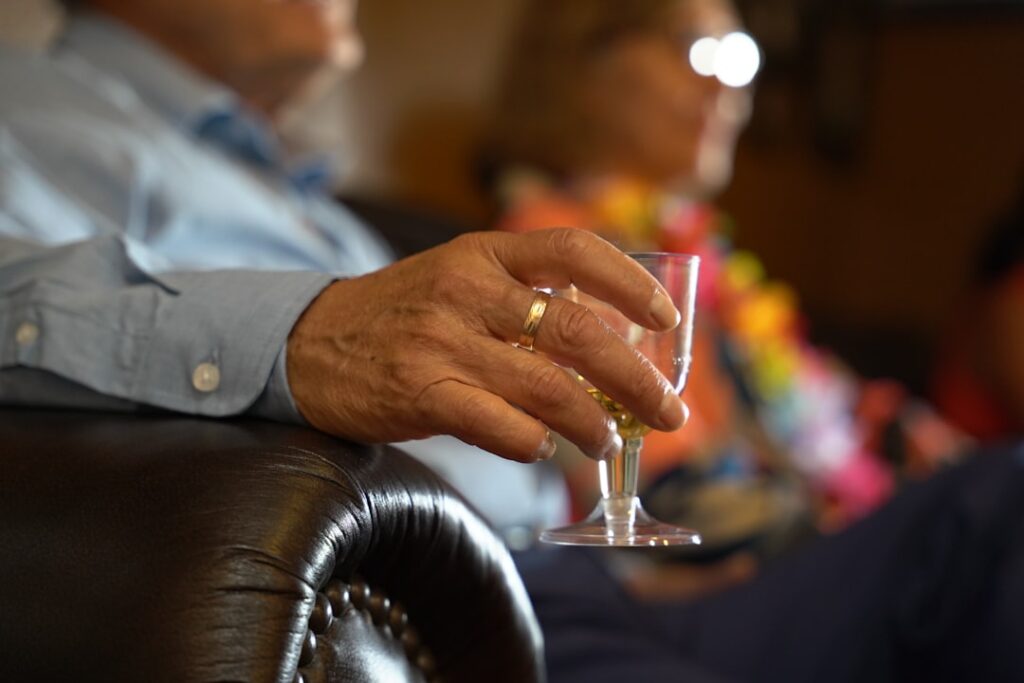 An elderly person wearing a gold ring, holding a small glass goblet.