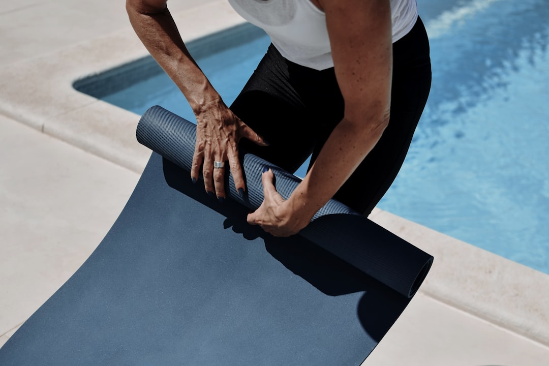 An elderly person rolling up a yoga mat, standing next to a swimming pool.