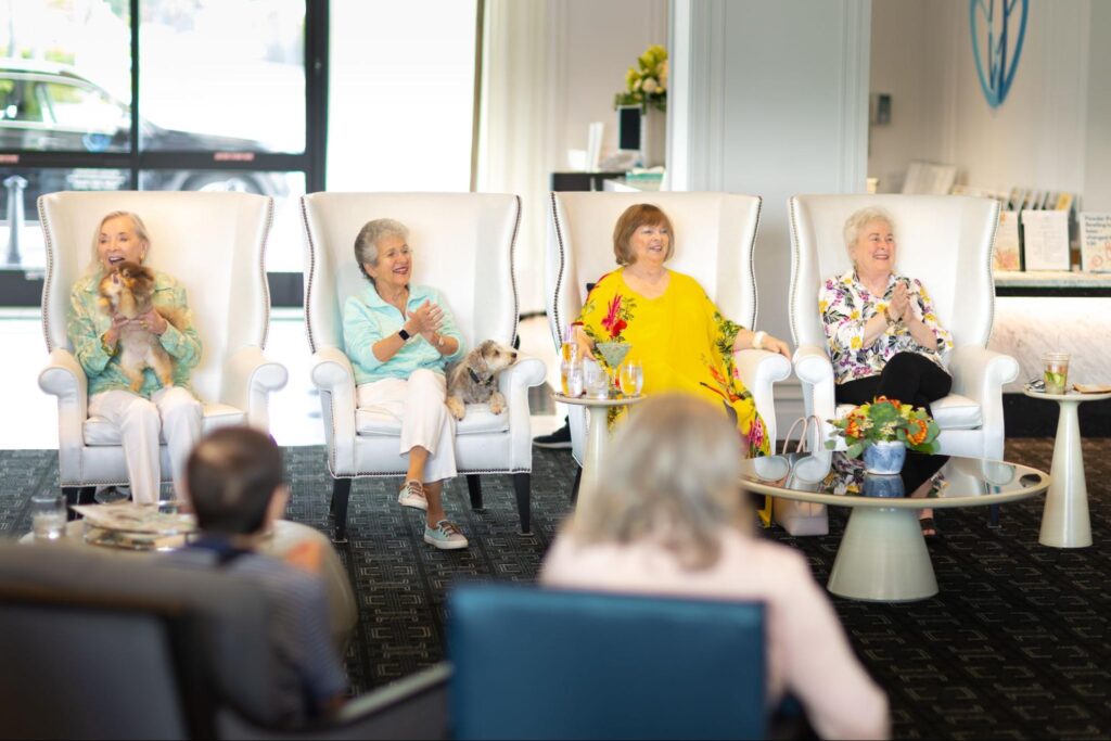 Four elderly women, sitting in a row of luxurious, white armchairs, smiling.