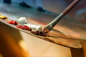 A paint brush being dipped into a plate containing different colored paints.
