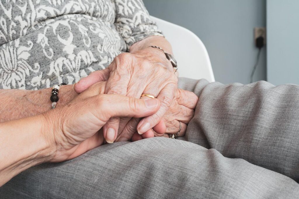 An elderly person holding their hands in their lap, holding another person’s hand.