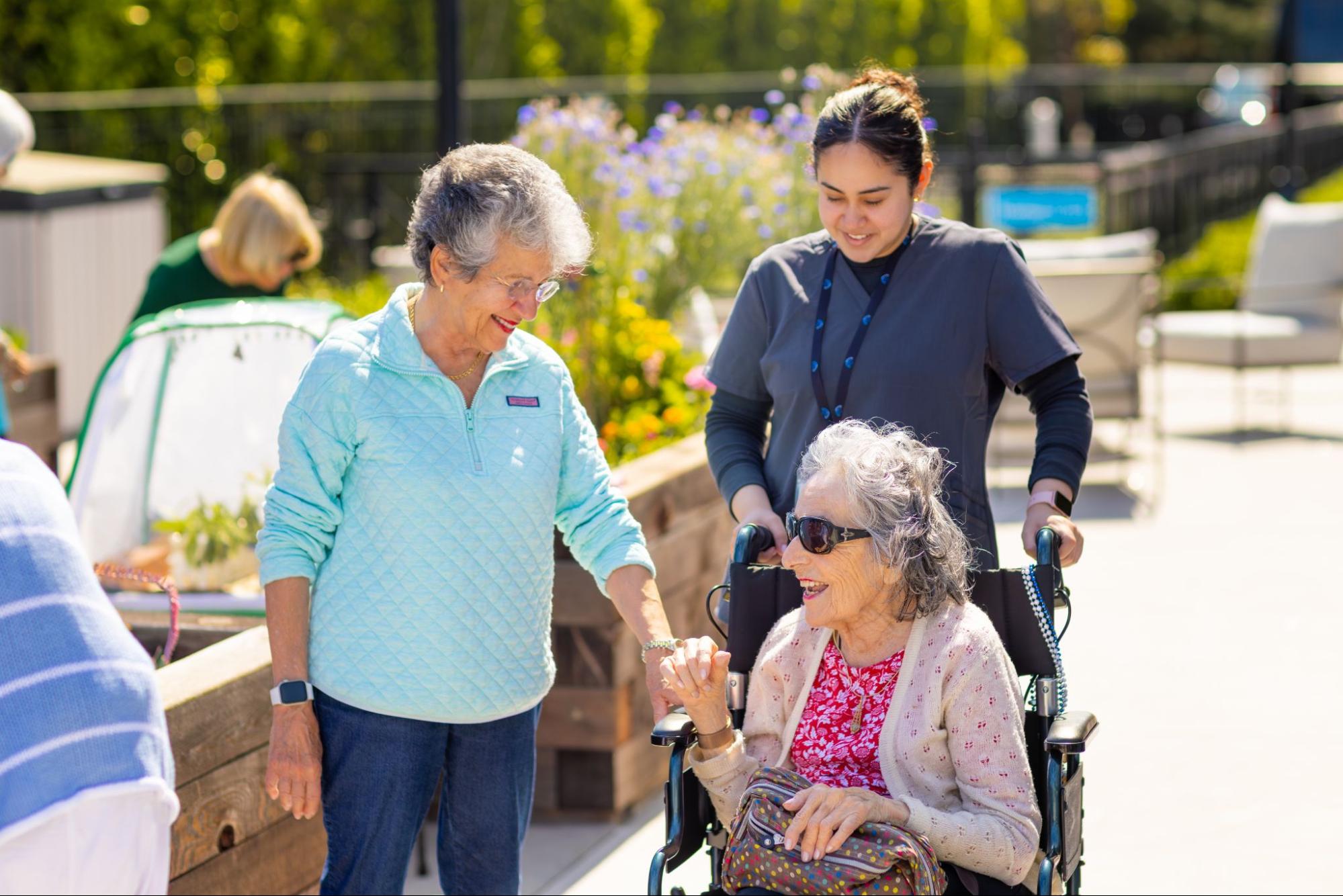 An assisted living coordinator pushing an elderly woman in a wheelchair outside, with another woman standing next to them and smiling.