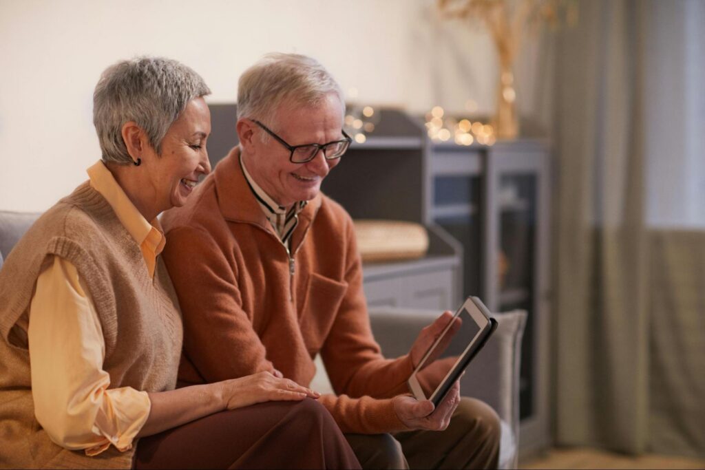 An elderly couple sitting together, looking at a tablet and smiling.