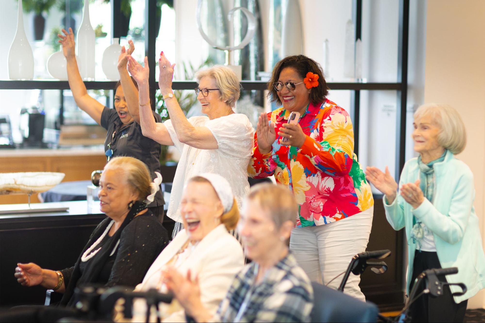 A group of senior women and staff members are smiling and clapping joyfully during a community event.