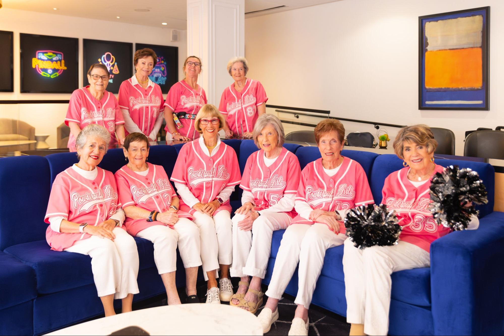 A group of ten senior women, some holding pom-poms, pose together in matching pink baseball jerseys while sitting on a blue sofa.