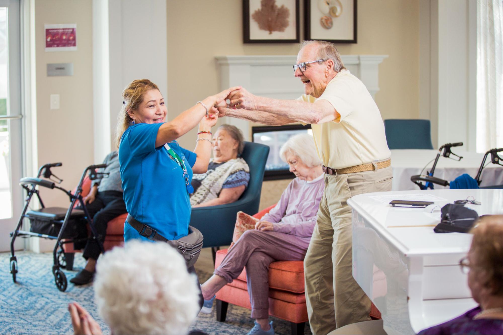 A smiling staff member in a blue shirt dances with a joyful senior man in a communal living room setting.