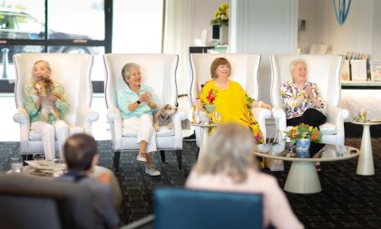 Four elderly women sitting in luxury, white arm chairs in a lounge, smiling.