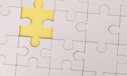 A close-up view of a white jigsaw puzzle with one piece missing, revealing a solid yellow background underneath.