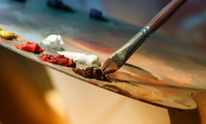 A paint brush being dipped into a plate containing different colored paints.